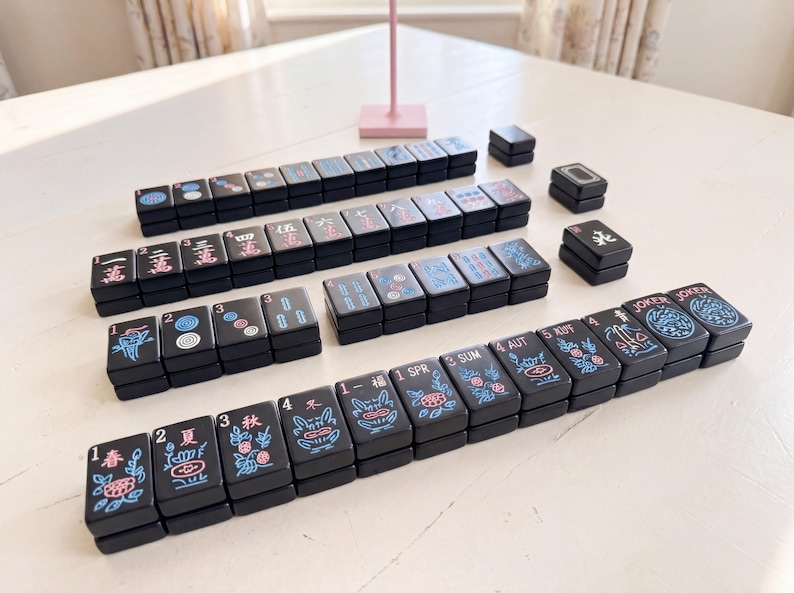 May include: A collection of black Mahjong tiles, displaying a variety of symbols and characters in red, blue, and white. The tiles are arranged on a white surface, demonstrating the game's layout. The set includes tiles with numbers, suits, and special characters.