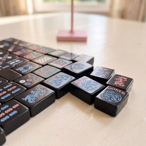 May include: A set of black Mahjong tiles with various symbols and characters in pink, blue, and white. Some tiles feature dragon and floral designs. The word "Joker" is visible on some tiles. The tiles are arranged on a light-coloured surface.