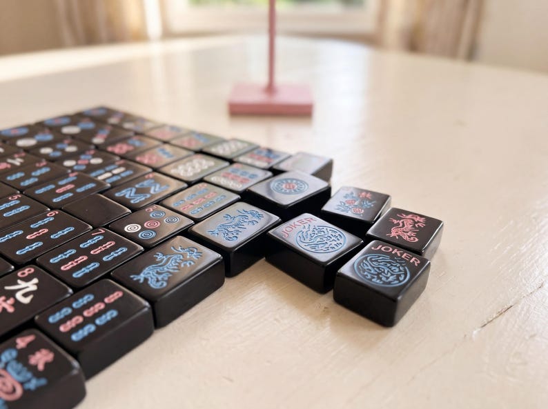 May include: A set of black Mahjong tiles with various symbols and characters in pink, blue, and white. Some tiles feature dragon and floral designs. The word "Joker" is visible on some tiles. The tiles are arranged on a light-coloured surface.