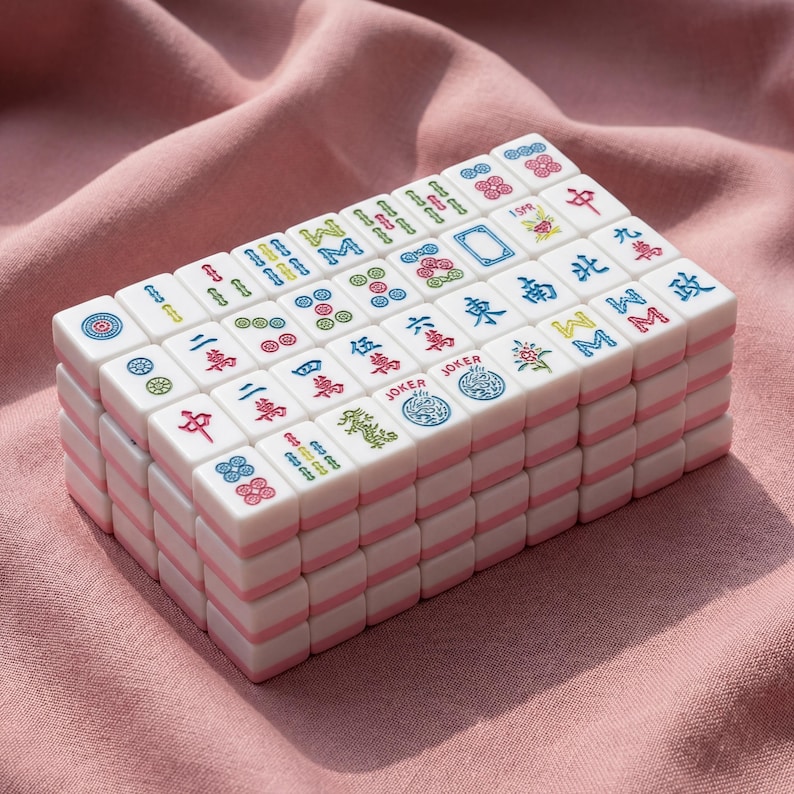 May include: A stack of white Mahjong tiles with pink edges, featuring colorful symbols and Chinese characters. The tiles are arranged neatly, showcasing various designs and colors, including red, green, blue, and yellow. The game pieces are set against a pink fabric background.