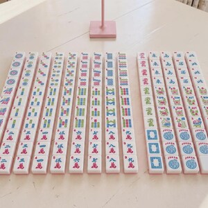 May include: A set of white mahjong tiles arranged in rows on a light surface. The tiles feature colourful engraved symbols, Chinese characters, numbers, and English text including Joker, Spr, Sum, Aut, and Win. One pink tile is at the end of a row.