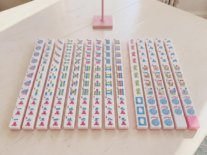May include: A set of white mahjong tiles arranged in rows on a light surface. The tiles feature colourful engraved symbols, Chinese characters, numbers, and English text including Joker, Spr, Sum, Aut, and Win. One pink tile is at the end of a row.