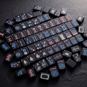 K&ouml;nnte beinhalten: Eine Sammlung schwarzer Mahjong-Steine mit roten, blauen und wei&szlig;en Symbolen und Schriftzeichen. Die Steine sind auf einer dunklen Oberfl&auml;che angeordnet und zeigen verschiedene Anz&uuml;ge, Zahlen und Spezialsteine wie "Joker".