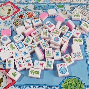 May include: A collection of white Mahjong tiles with pink backs, featuring various green, blue, and red engraved symbols, numbers, and characters, scattered across a light blue fabric surface decorated with illustrated food and floral patterns.