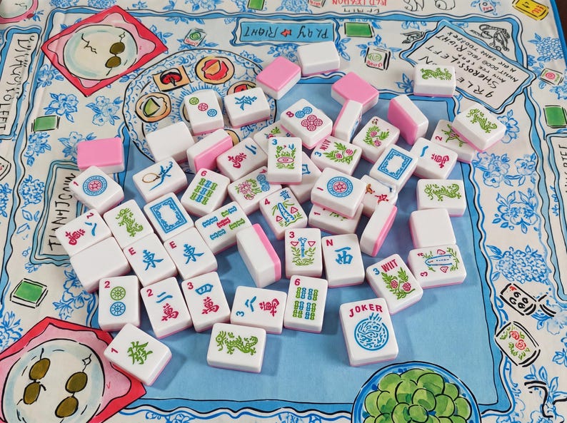 May include: A collection of white Mahjong tiles with pink backs, featuring various green, blue, and red engraved symbols, numbers, and characters, scattered across a light blue fabric surface decorated with illustrated food and floral patterns.