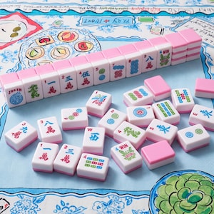 May include: A set of Mahjong tiles featuring a white face with colorful Chinese characters, symbols, and numbers, and a solid pink back. The tiles are arranged in a neat row and scattered across a light blue and white patterned fabric surface.