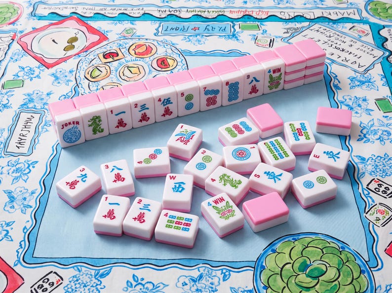 May include: A set of Mahjong tiles featuring a white face with colorful Chinese characters, symbols, and numbers, and a solid pink back. The tiles are arranged in a neat row and scattered across a light blue and white patterned fabric surface.