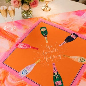 May include: An orange and pink decorative table runner with illustrations of champagne bottles. The runner has the words "Sip, Sparkle, Mahjong" in white script. The runner is surrounded by a pink border.