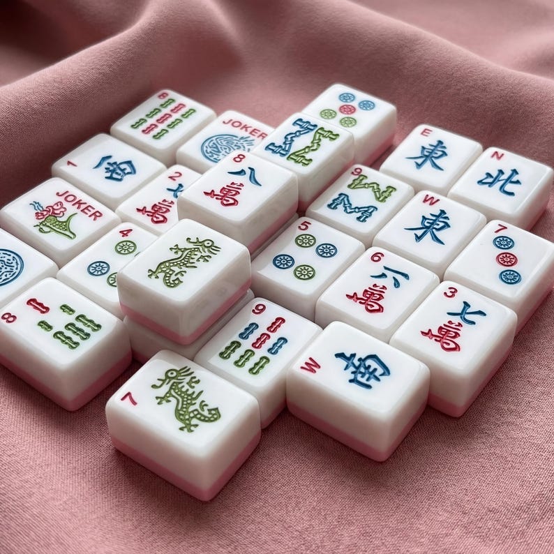 May include: A collection of white and pink Mahjong tiles with various symbols and characters in red, green, and blue. The tiles are arranged on a pink fabric surface, showcasing the game's components.