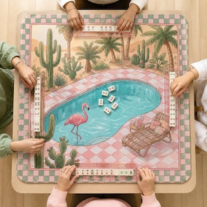 May include: A square mahjong mat featuring a desert oasis illustration with a swimming pool, a pink flamingo, cacti, palm trees, and a lounge chair. The border has a pink and green checkered pattern. Four clear tile racks hold mahjong tiles.