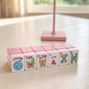 May include: A set of white and pink Mahjong tiles with various symbols and numbers. The tiles are arranged on a white surface, with a pink stand in the background. The tiles include the words "JOKER", "SPR", and "SUM".