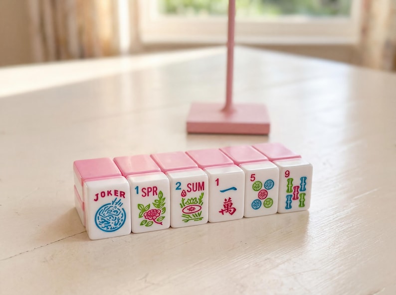 May include: A set of white and pink Mahjong tiles with various symbols and numbers. The tiles are arranged on a white surface, with a pink stand in the background. The tiles include the words "JOKER", "SPR", and "SUM".