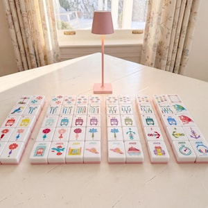 May include: A set of white mahjong tiles with colorful graphic illustrations including flowers, travel icons, and animals, arranged in rows on a white surface. A small, solid pink table lamp with a conical shade stands behind the tiles.