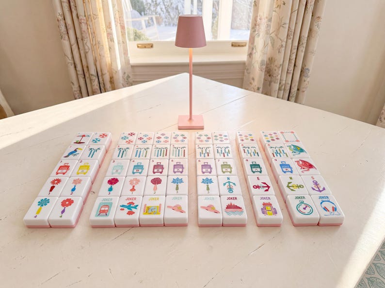 May include: A set of white mahjong tiles with colorful graphic illustrations including flowers, travel icons, and animals, arranged in rows on a white surface. A small, solid pink table lamp with a conical shade stands behind the tiles.