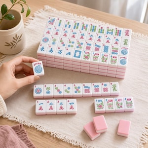 Może przedstawiać: Zestaw białych płytek do gry w Mahjong z różowymi spodami, ozdobionych kolorowymi chińskimi znakami, cyframi i symbolami w kolorze czerwonym, niebieskim i zielonym. Zestaw zawiera kolory, wiatry, smoki, kwiaty, pory roku oraz płytki Joker.