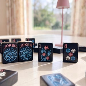 May include: A selection of black Mahjong tiles, each displaying different symbols and numbers, are arranged on a light surface. Two tiles read "JOKER" with a blue dragon design. Other tiles show the number "1" with floral designs and the number "8" with circles.