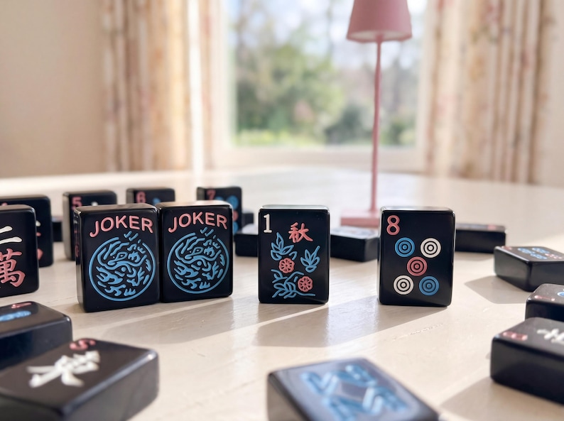 May include: A selection of black Mahjong tiles, each displaying different symbols and numbers, are arranged on a light surface. Two tiles read "JOKER" with a blue dragon design. Other tiles show the number "1" with floral designs and the number "8" with circles.