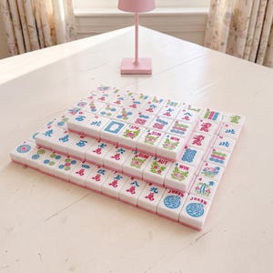 May include: A set of Mahjong tiles arranged in three layers on a white table. The tiles are white with pink edges and feature colorful symbols and characters. A pink lamp is in the background.