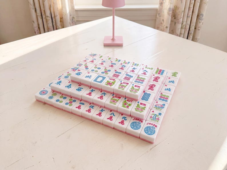 May include: A set of Mahjong tiles arranged in three layers on a white table. The tiles are white with pink edges and feature colorful symbols and characters. A pink lamp is in the background.