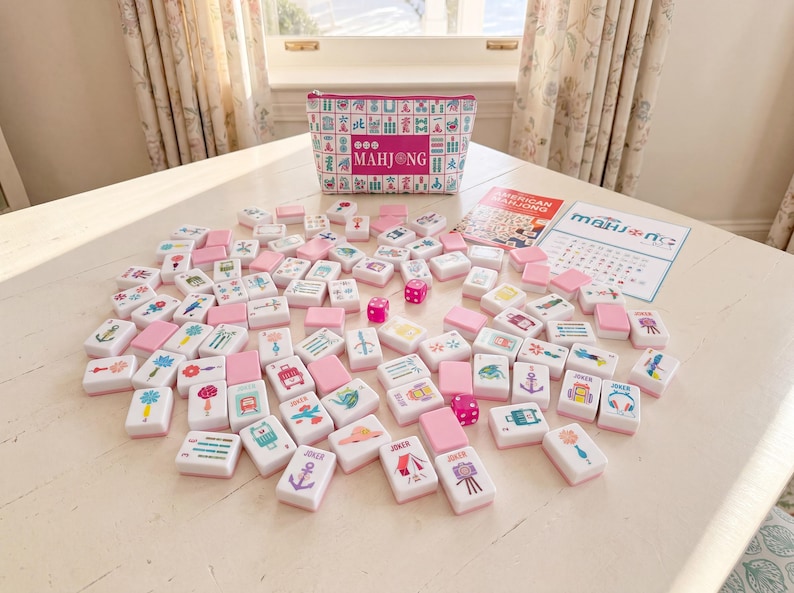 May include: A Mahjong game set featuring white tiles with colorful illustrations and pink backs, a matching zippered storage pouch with a Mahjong pattern, three pink dice, and two instructional cards with the text American Mahjong and Mahjong.