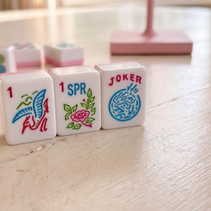 May include: White Mahjong tiles with pink accents and colorful illustrations. The tiles feature images of a bird, flowers, and the word "JOKER". The game pieces are arranged on a light-colored wooden surface.