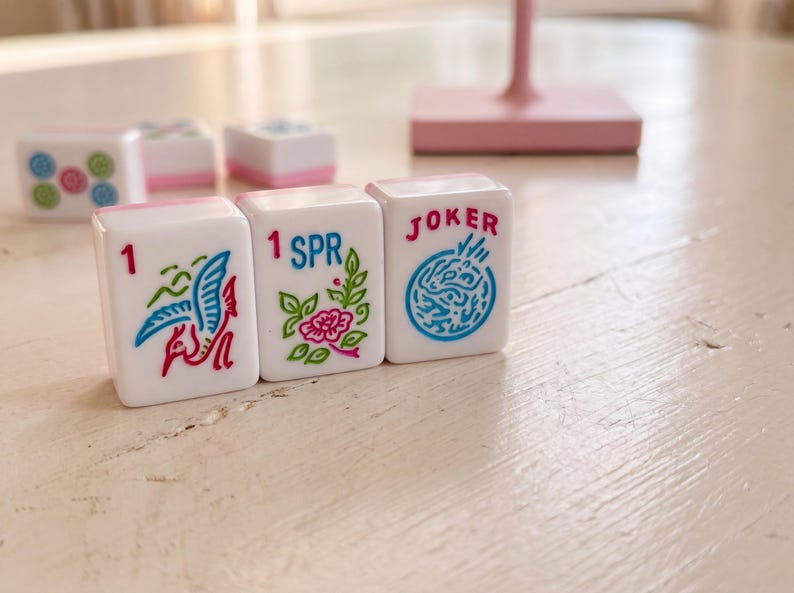 May include: White Mahjong tiles with pink accents and colorful illustrations. The tiles feature images of a bird, flowers, and the word "JOKER". The game pieces are arranged on a light-colored wooden surface.