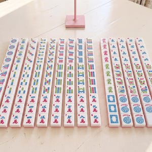 May include: A complete set of Mahjong tiles arranged on a light-colored surface. The tiles are white with pink, blue, green, and red markings. The set includes tiles with numbers, suits, and characters, along with joker tiles.