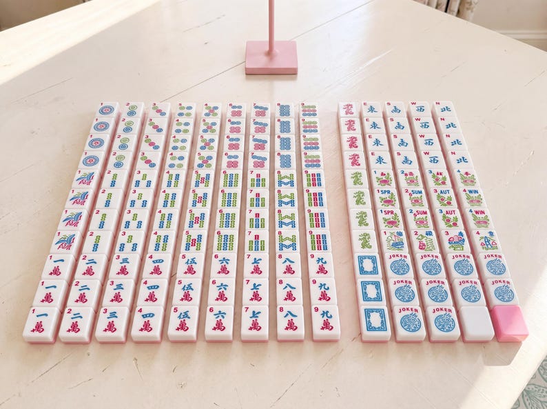 May include: A complete set of Mahjong tiles arranged on a light-colored surface. The tiles are white with pink, blue, green, and red markings. The set includes tiles with numbers, suits, and characters, along with joker tiles.