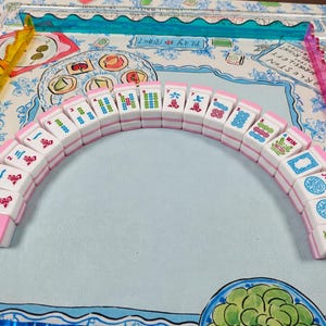 May include: A set of white and pink Mahjong tiles arranged in a semi-circle, featuring various colorful Chinese characters, numbers, and symbols including Joker tiles. The tiles are positioned on a game mat with illustrated food and text.