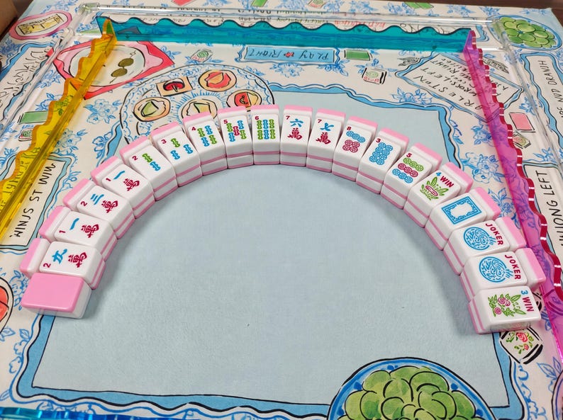 May include: A set of white and pink Mahjong tiles arranged in a semi-circle, featuring various colorful Chinese characters, numbers, and symbols including Joker tiles. The tiles are positioned on a game mat with illustrated food and text.