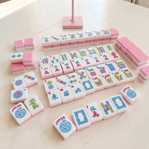 May include: A complete Mahjong set with white tiles and pink accents. The tiles display various symbols, numbers, and Chinese characters. The set includes tiles labeled "JOKER", "SPR", "SUM", and "AUT". The game is arranged on a white surface.