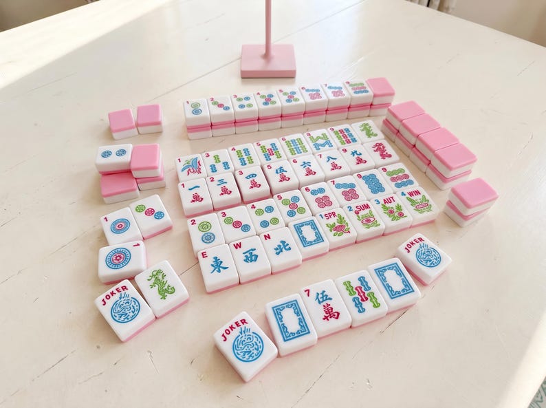 May include: A complete Mahjong set with white tiles and pink accents. The tiles display various symbols, numbers, and Chinese characters. The set includes tiles labeled "JOKER", "SPR", "SUM", and "AUT". The game is arranged on a white surface.