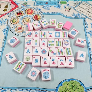 May include: A set of white and pink Mahjong tiles with colorful symbols, numbers, and characters arranged on a light blue fabric mat featuring illustrated food items and the text PLAY RIGHT and WRLESTON.