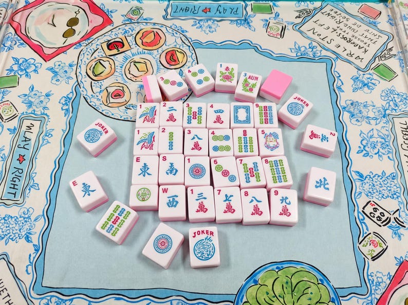 May include: A set of white and pink Mahjong tiles with colorful symbols, numbers, and characters arranged on a light blue fabric mat featuring illustrated food items and the text PLAY RIGHT and WRLESTON.