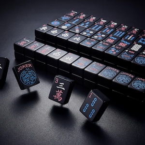 K&ouml;nnte beinhalten: Ein schwarzes Mahjong-Set mit Spielsteinen, die chinesische Schriftzeichen und Symbole zeigen. Einige Spielsteine zeigen das Wort "JOKER" und dekorative Designs in Blau und Rot. Das Set ist auf einer dunklen Oberfl&auml;che angeordnet.