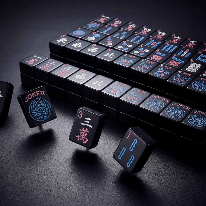 K&ouml;nnte beinhalten: Ein schwarzes Mahjong-Set mit Spielsteinen, die chinesische Schriftzeichen und Symbole zeigen. Einige Spielsteine zeigen das Wort "JOKER" und dekorative Designs in Blau und Rot. Das Set ist auf einer dunklen Oberfl&auml;che angeordnet.