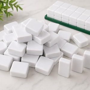May include: A collection of white Mahjong tiles, some scattered and others neatly arranged in a green tray. The tiles are square with rounded edges, suggesting a classic game set. Green foliage is visible in the background.