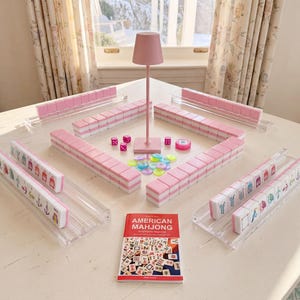 May include: A Mahjong set featuring pink and white tiles arranged on clear acrylic racks, a pink central lamp, colorful translucent betting chips, pink dice, and a red instructional booklet titled AMERICAN MAHJONG Simplified for Beginners.