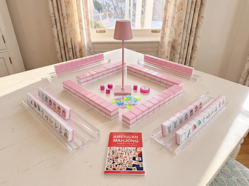 May include: A Mahjong set featuring pink and white tiles arranged on clear acrylic racks, a pink central lamp, colorful translucent betting chips, pink dice, and a red instructional booklet titled AMERICAN MAHJONG Simplified for Beginners.