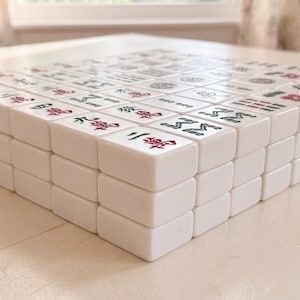 May include: A close-up of a Mahjong set with white tiles. The tiles are stacked, revealing the red and green symbols and characters. The game is set on a white surface, with a window in the background.