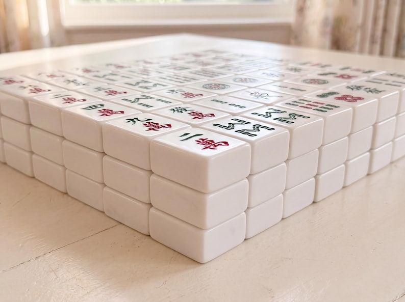 May include: A close-up of a Mahjong set with white tiles. The tiles are stacked, revealing the red and green symbols and characters. The game is set on a white surface, with a window in the background.