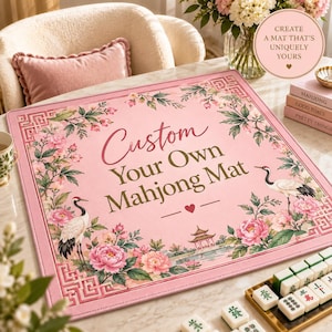 Może przedstawiać: Kwadratowa różowa mata do mahjonga z kwiatową obwódką z piwonii, zielonych liści i dwóch żurawi. Środkowy tekst brzmi Custom Your Own Mahjong Mat z małym sercem. Rogi mają geometryczny wzór, a na dole znajduje się ilustracja pagody.