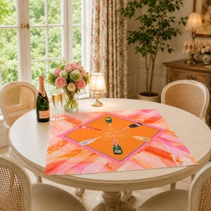 May include: A round white table set for a celebration. A pink and orange patterned table runner with champagne bottle illustrations and the text "Sip Sparkle Mahjong" is centered on the table. A bottle of champagne, two glasses, and a bouquet of flowers are also on the table.