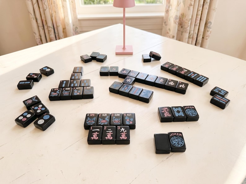 May include: Black Mahjong tiles with red, blue, and white symbols and characters are arranged on a white surface. The tiles are scattered, suggesting a game. A pink lamp and a window are visible in the background. The game pieces are in various arrangements.