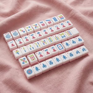 May include: A collection of white Mahjong tiles with red, blue, and green markings, including Chinese characters and numbers. The tiles are arranged on a pink fabric surface, showcasing the game's components.