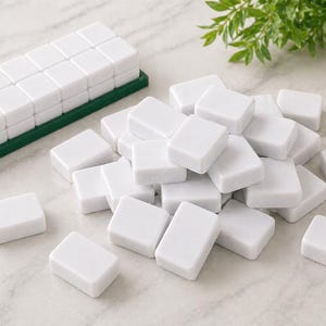 May include: A collection of white rectangular tiles, some scattered and others neatly stacked on a green tray. The tiles are smooth and uniform, suggesting a game or puzzle. The background includes greenery and a decorative vase.