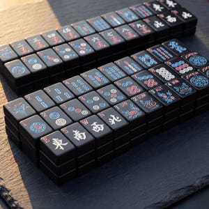 K&ouml;nnte beinhalten: Zwei S&auml;tze schwarzer Mahjong-Steine sind ordentlich gestapelt und zeigen verschiedene Symbole und Zahlen in Rot, Blau und Wei&szlig;. Die Steine sind auf einer dunklen Oberfl&auml;che angeordnet und zeigen die komplizierten Designs und Komponenten des Spiels.