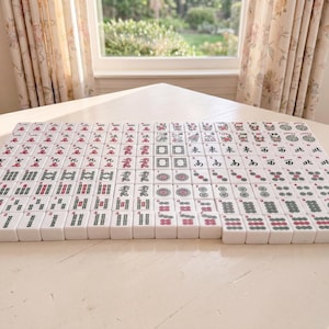May include: A complete set of Mahjong tiles arranged on a light-colored surface. The tiles are white with red, green, and black symbols and characters. The game set is displayed in front of a window with floral curtains.