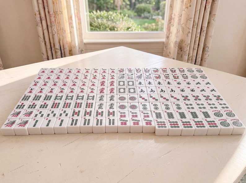 May include: A complete set of Mahjong tiles arranged on a light-colored surface. The tiles are white with red, green, and black symbols and characters. The game set is displayed in front of a window with floral curtains.