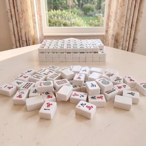 May include: A complete Mahjong set with white tiles featuring red and green symbols and characters. The tiles are scattered on a light-colored surface, with a stack of tiles in the background. The game is set up on a table near a window.